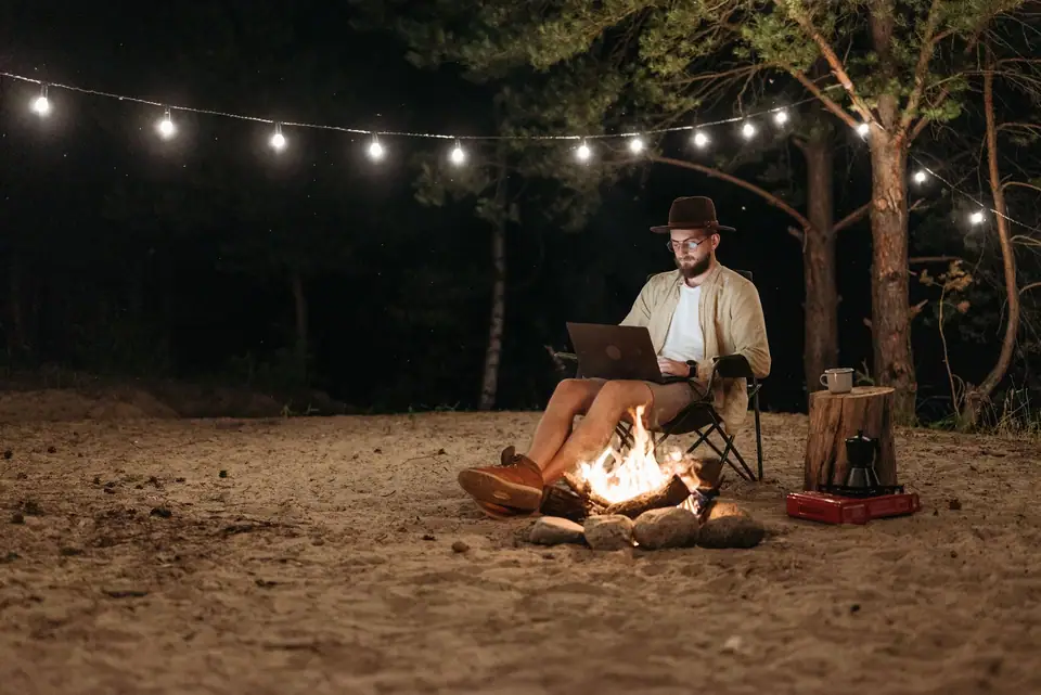 A man working on a laptop by a campfire under string lights at a forest campsite